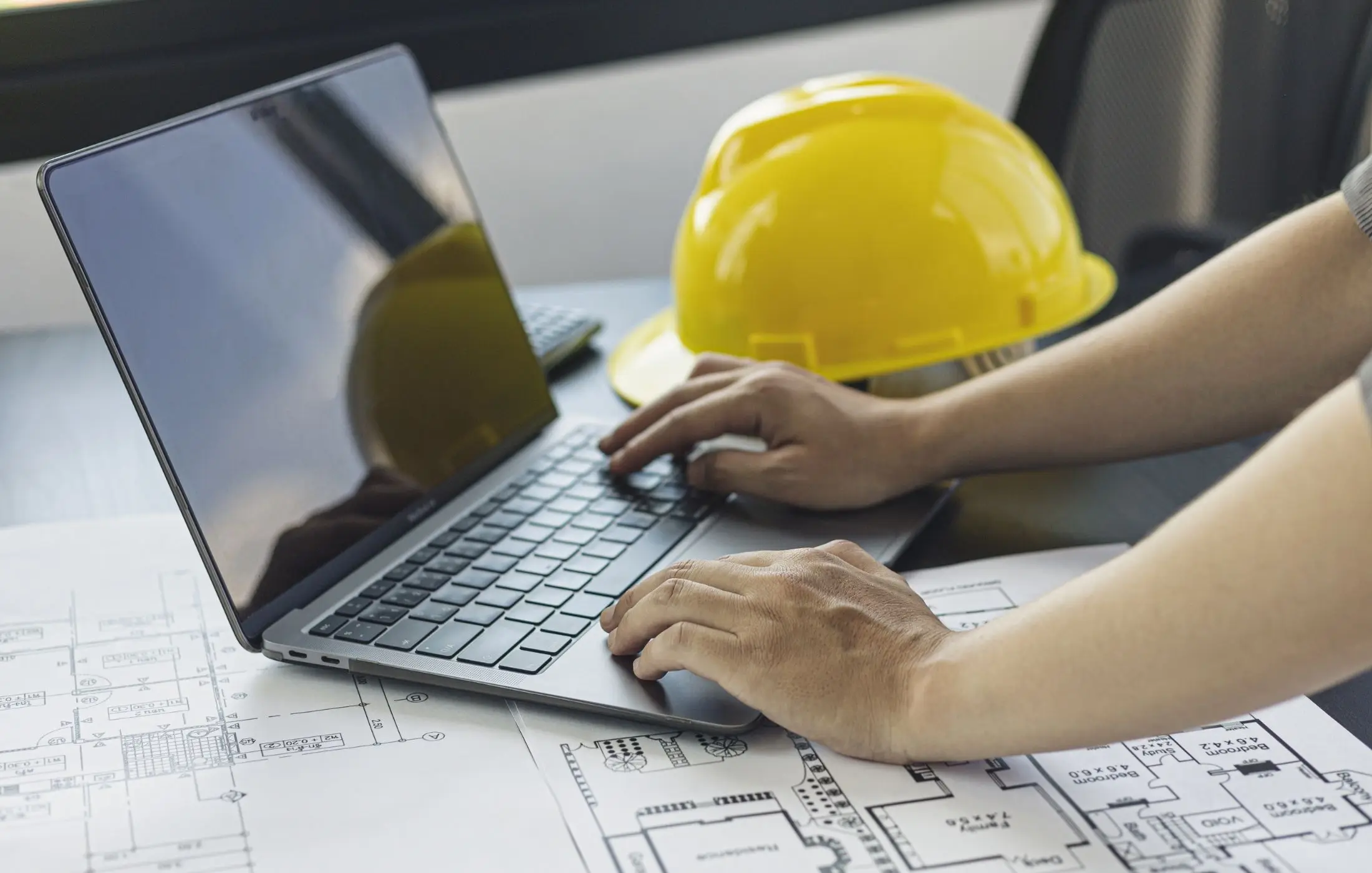 Engineer typing on a laptop next to a yellow hard hat and construction plans.