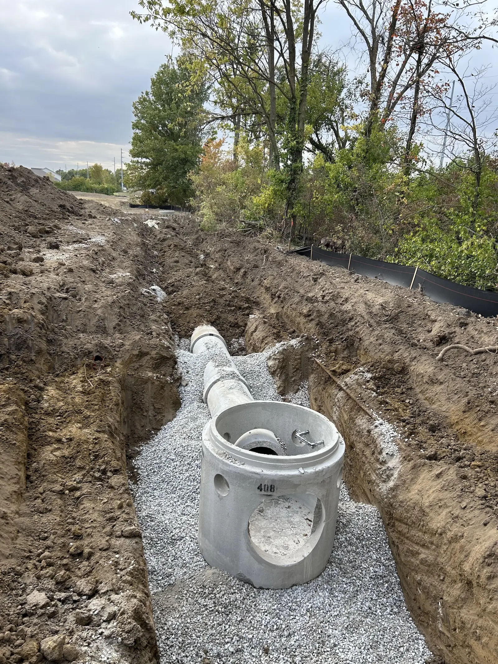 Concrete utility manhole and pipe installed in a gravel bed within a dirt trench.